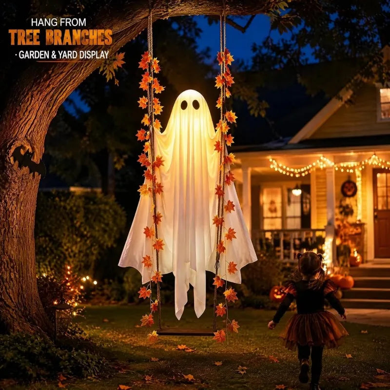 A glowing Halloween ghost yard decoration hanging from a tree swing on a lawn in front of a house at night. The ghost has a white sheet-like body with black eyes and mouth, decorated with orange and red autumn leaves.