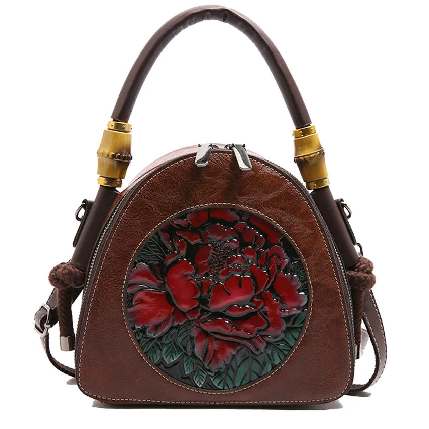 A brown faux leather handbag with a top handle and shoulder strap, featuring a circular 3D floral design on the front in red and green, with gold-colored metal accents on the handle attachments.